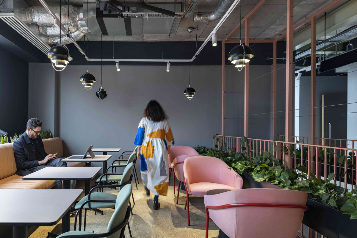 A dining space in the office. The chairs on the right of the space are light pink, while the walls, tables and seating on the left are in dark, muted colours. A line on plants is positioned behind the pink chairs.